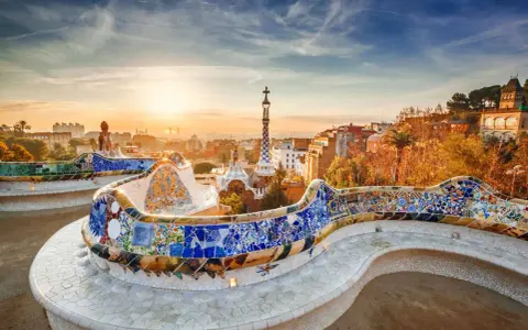 Bancos de mosaico coloridos en el Park Güell, un lugar imprescindible que ver en Barcelona, con la ciudad al amanecer.