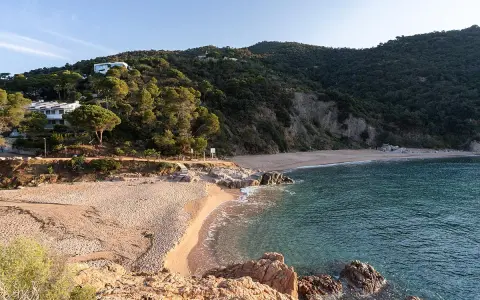 La platja de Canyerets, con su arena dorada y aguas cristalinas, se extiende bajo una colina verde y casas blancas.
