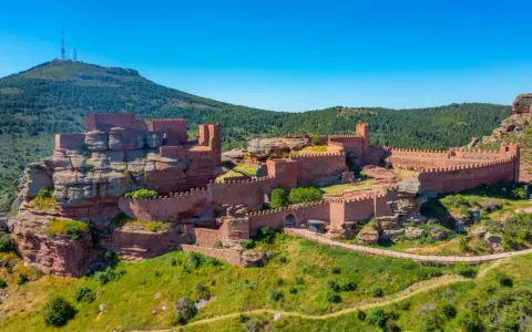 Imponente castillo de Peracense, una fortaleza de piedra rojiza que se alza sobre un peñón, rodeada de vegetación y bajo un cielo azul intenso.
