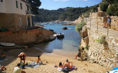 Gente relajándose en la arena de cala Malaret, Begur, con barcos anclados en aguas cristalinas y colinas verdes al fondo.