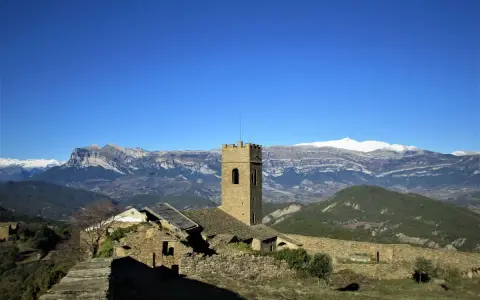 Antiguo muro de roda con torreón de piedra y ruinas, rodeado de montañas nevadas bajo un cielo azul intenso.