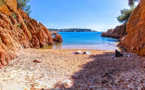 Arena dorada y aguas turquesas en la cala Maset. Dos personas toman el sol tranquilamente en la playa, rodeadas de imponentes rocas rojizas.