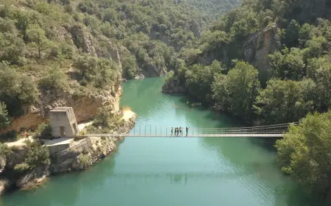 Un puente colgante cruza un río turquesa rodeado de montañas verdes. Un grupo de personas camina por el puente de Montañana.