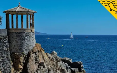 Un mirador de piedra con columnas se alza sobre rocas, con el mar azul y veleros al fondo. Un lugar perfecto para pasear por los **caminos de ronda**.