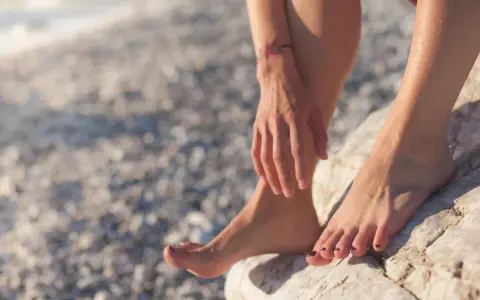 Pies descalzos sobre rocas en una playa nudista de Cadaqués. El sol brilla sobre la piel mojada y un pequeño tatuaje de flores adorna el brazo.