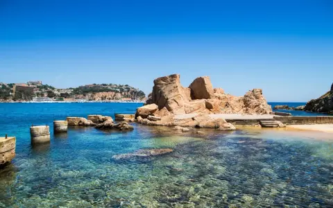 Un paseo marítimo en Sant Feliu de Guíxols, con rocas doradas y aguas cristalinas. El pueblo se alza en la colina.