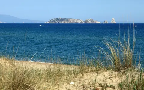 Dunas de hierba seca en primer plano, mar azul y un archipiélago rocoso en la distancia, evocando la belleza de la playa mas Pinell.