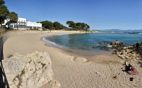 Platja Cala de la Creu: arena dorada, aguas turquesas y un hotel blanco con vistas al mar. Familias disfrutan del sol y juegan en la arena.