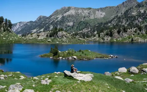 Persona sentada en roca contempla los lagos de Colomers en una ruta corta. Paisaje montañoso y azul intenso.