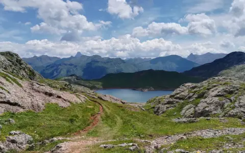 Sendero de montaña que conduce al sereno ibon de Estanes, rodeado de picos escarpados y un cielo azul con nubes algodonosas.