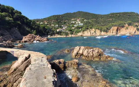 Un puente de piedra cruza aguas turquesas en la cala Canyet. Barcas flotan tranquilas mientras colinas verdes y casas blancas coronan el paisaje.
