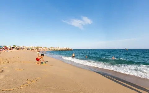 Un niño juega en la arena dorada de la playa de les barques, mientras otros disfrutan del mar azul y un velero navega en el horizonte.