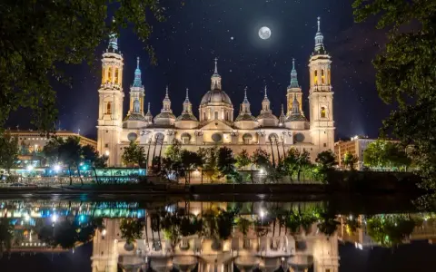 La majestuosa basílica del Pilar, iluminada bajo un cielo estrellado y la luna, se refleja en el río.