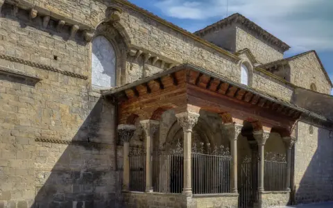 La **catedral de Jaca**, con su imponente arquitectura románica, se alza bajo un cielo azul. Un pórtico de madera y piedra protege la entrada.
