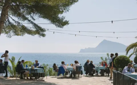 Gente disfrutando de una comida al aire libre con vistas al mar. Un lugar ideal donde comer en Altea, con mesas azules y un ambiente relajado bajo un árbol.