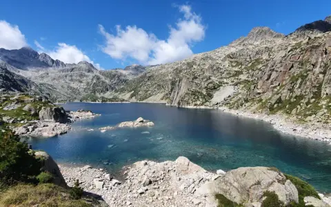 Lago de aguas cristalinas rodeado de montañas rocosas en el Vall de Boí. El cielo azul con nubes blancas contrasta con el verde de la vegetación.