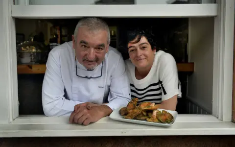 Chef y camarera sonríen tras ventana con plato de setas. Descubre dónde comer en Castro Urdiales.