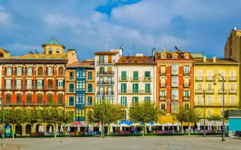 Fachada de edificios coloridos en una plaza, ideal para saber **qué ver en Pamplona**. Terrazas y árboles verdes bajo un cielo azul.