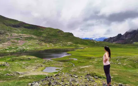 Mujer contempla los ibones del Valle de Tena, un paisaje de lagos y prados verdes bajo un cielo nublado.