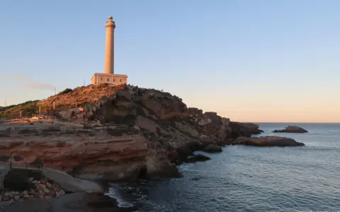 Faro sobre acantilados rojizos, con el mar azul y un cielo despejado. Un camino desciende hacia una pequeña cala roja.