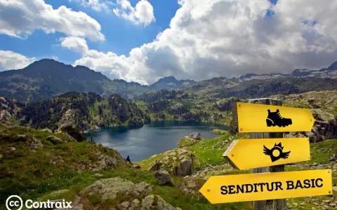 Paisaje de montaña con un lago azul, señalización de sendero y el letrero "SENDITUR BASIC", parte de la ruta de los 7 lagos Pirineos.