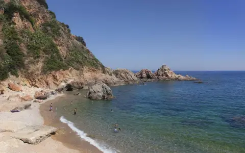 Les platges de Blanes ofereixen aigües cristal·lines i roques imponents sota un cel blau. Gent gaudeix del sol i del mar.