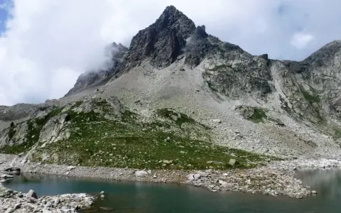 Paisaje montañoso con un lago de aguas tranquilas en primer plano. Las laderas están cubiertas de rocas y vegetación verde, con picos escarpados y nubes en el cielo. Los **ibones de Arriel** reflejan la majestuosidad del entorno.