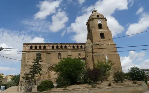 Iglesia de piedra con torre y reloj, rodeada de vegetación. Un día soleado en Alberuela de Tubo.