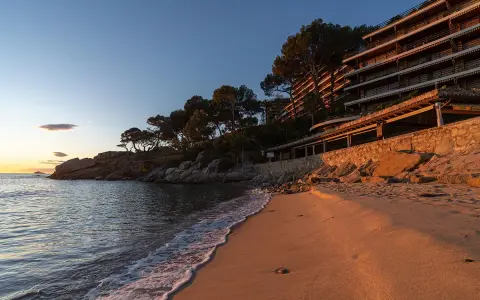 Atardecer dorado en la playa, con olas suaves lamiendo la arena. Al fondo, la silueta de la playa torre valentina se alza entre pinos.