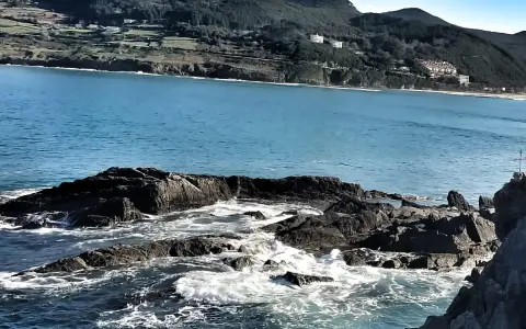 Olas rompiendo en rocas oscuras, con la costa verde y edificios de Mundaka al fondo. Un lugar perfecto para comer y disfrutar del paisaje.