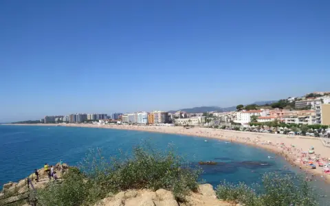Vista panorámica de la playa S'Abanell en Blanes, con el mar azul intenso, arena dorada repleta de gente y edificios costeros bajo un cielo despejado.