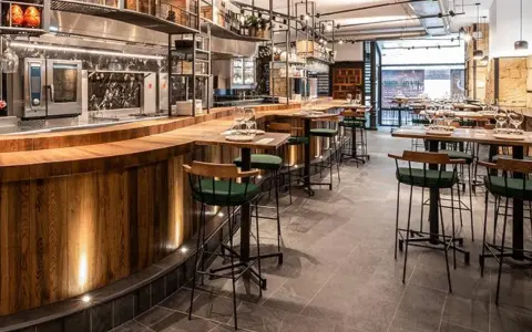 Bar moderno con taburetes verdes y mesas de madera. Un lugar ideal donde comer en Salamanca, con cocina a la vista y ambiente acogedor.