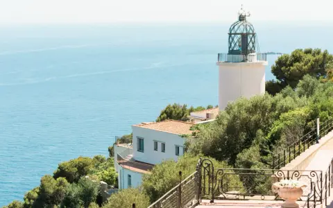 El faro de Sant Sebastià se alza sobre el mar azul, rodeado de vegetación exuberante.