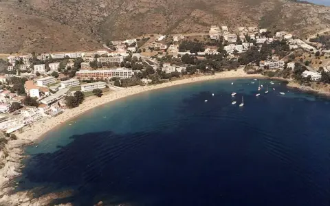 Vista aérea de la playa Almadrava en Roses, con edificios blancos y barcos anclados en aguas azules.