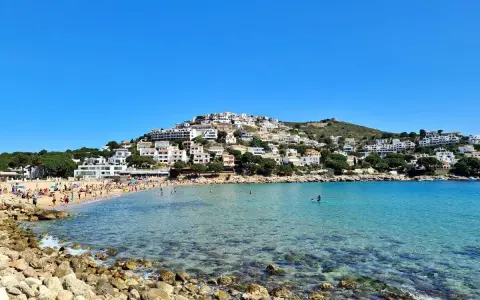 Vista de la platja de Montgó, con gente disfrutando del sol y el mar turquesa, y casas blancas escalonadas en la colina.