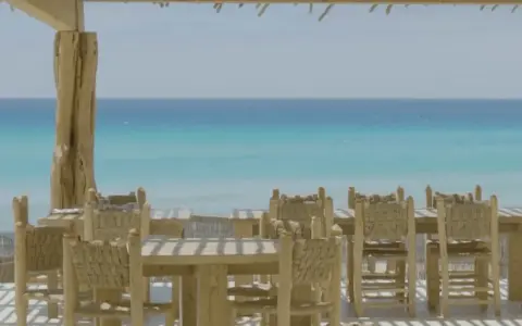 Mesas y sillas de madera rústica en la terraza de restaurantes en Formentera, con el mar turquesa de fondo.