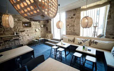Ambiente acogedor para comer en Allariz. Mesas de madera, bancos con cojines y lámparas de mimbre en un local de piedra.