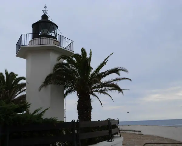 El faro de Palamós, blanco y esbelto, se alza junto a palmeras y el mar azul.