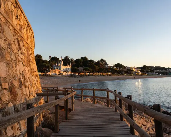Pasarela de madera hacia la platja Sant Pol, Sant Feliu de Guíxols. Edificios blancos y palmeras bordean la arena dorada bajo un cielo azul.
