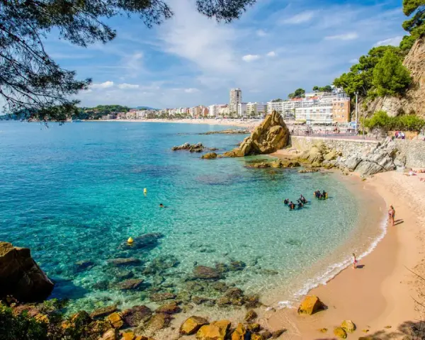 Una caleta en la Costa Brava con aguas cristalinas, buceadores explorando el fondo rocoso y una playa con gente disfrutando del sol.