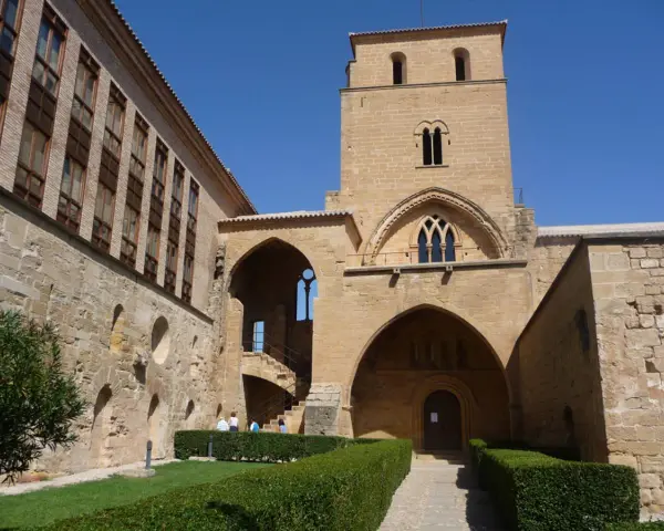 El **castillo de Alcañiz**, con su imponente torre y arcos, se alza bajo un cielo azul. Jardines cuidados y muros de piedra evocan su historia.