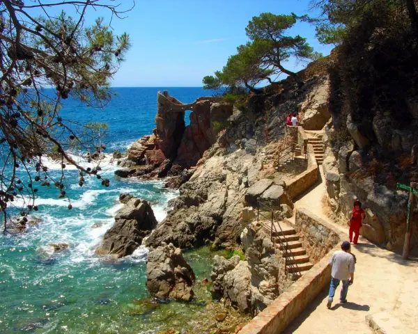Un camino de ronda con escaleras de piedra serpentea por la costa rocosa. El mar turquesa rompe contra las rocas.