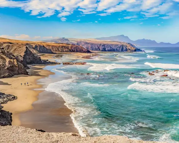 Paisaje de las playas de Fuerteventura con acantilados rocosos, arena dorada y el mar turquesa con olas rompiendo.