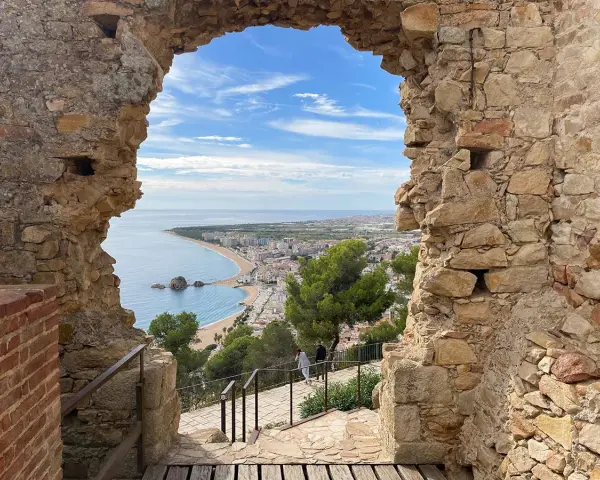 Vista desde el **castillo de Blanes** hacia la bahía y la ciudad. El mar azul contrasta con las ruinas de piedra y el cielo nublado.