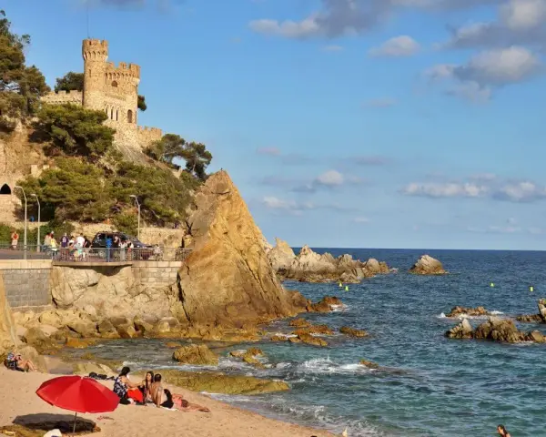 Castillo medieval sobre acantilados rocosos en sa caleta Lloret de Mar. Playa con gente y mar azul.