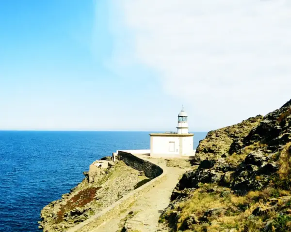 Un faro blanco se alza sobre un acantilado rocoso, vigilando el mar azul. Un camino serpentea hacia él, invitando a explorar los **faros Costa Brava**.