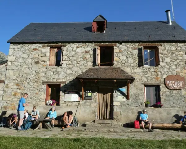 Grupo de excursionistas descansa frente al Refugio Viados, parte de la ruta de los tres refugios. Montañas y cielo azul al fondo.