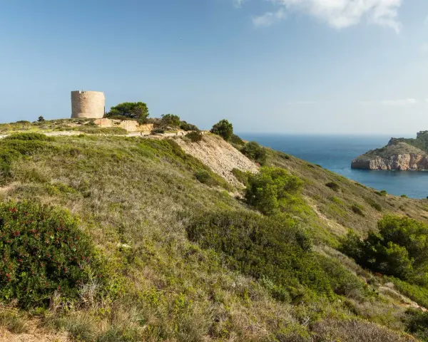 La torre de Montgó se alza sobre una colina verde, vigilando el mar azul y una bahía rocosa.