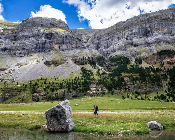 Senderista camina por un prado verde con imponentes montañas de **3000 del Pirineo** al fondo.
