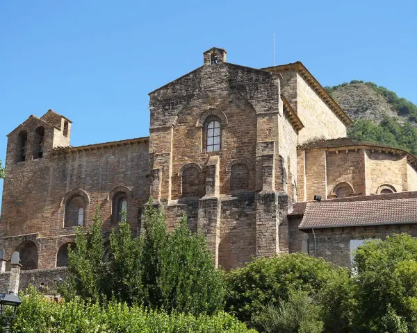 El **monasterio de Siresa**, con su imponente arquitectura de piedra y campanario, se alza bajo un cielo azul, rodeado de vegetación exuberante.